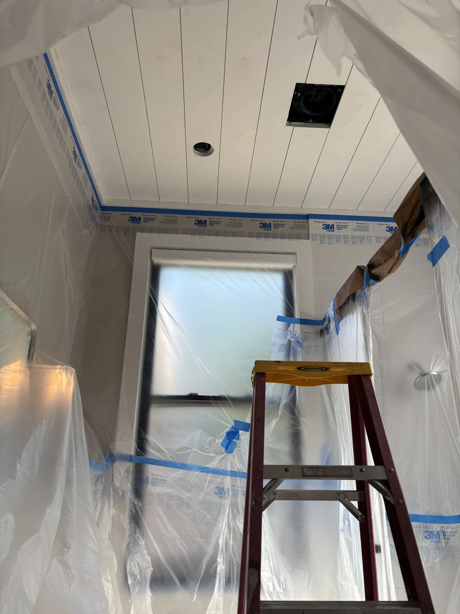 White plank bathroom ceiling above walls, a window and a shower wrapped in plastic masking film, with a stepladder in front
