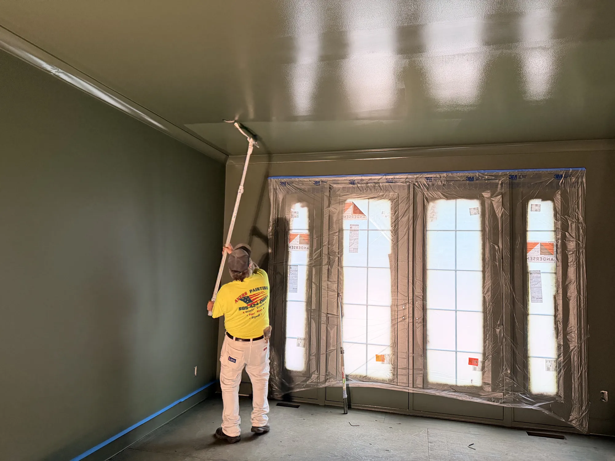 Painter seen from behind rolls a high-gloss ceiling with a long pole in a deep green room facing plastic-masked windows.