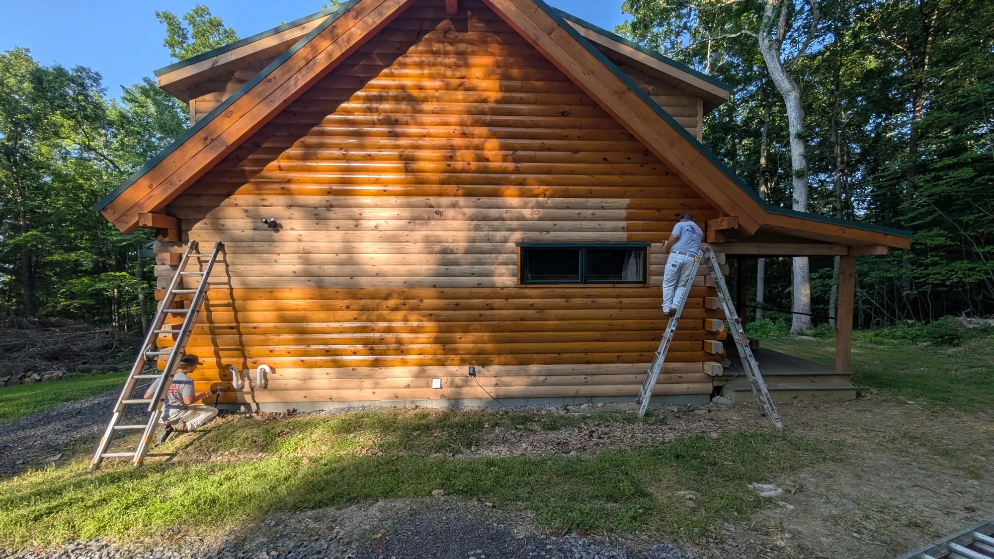 One painter on a stepladder and another crouched at the foot of an extension ladder apply finish to a sunlit log gable wall with tree shadows