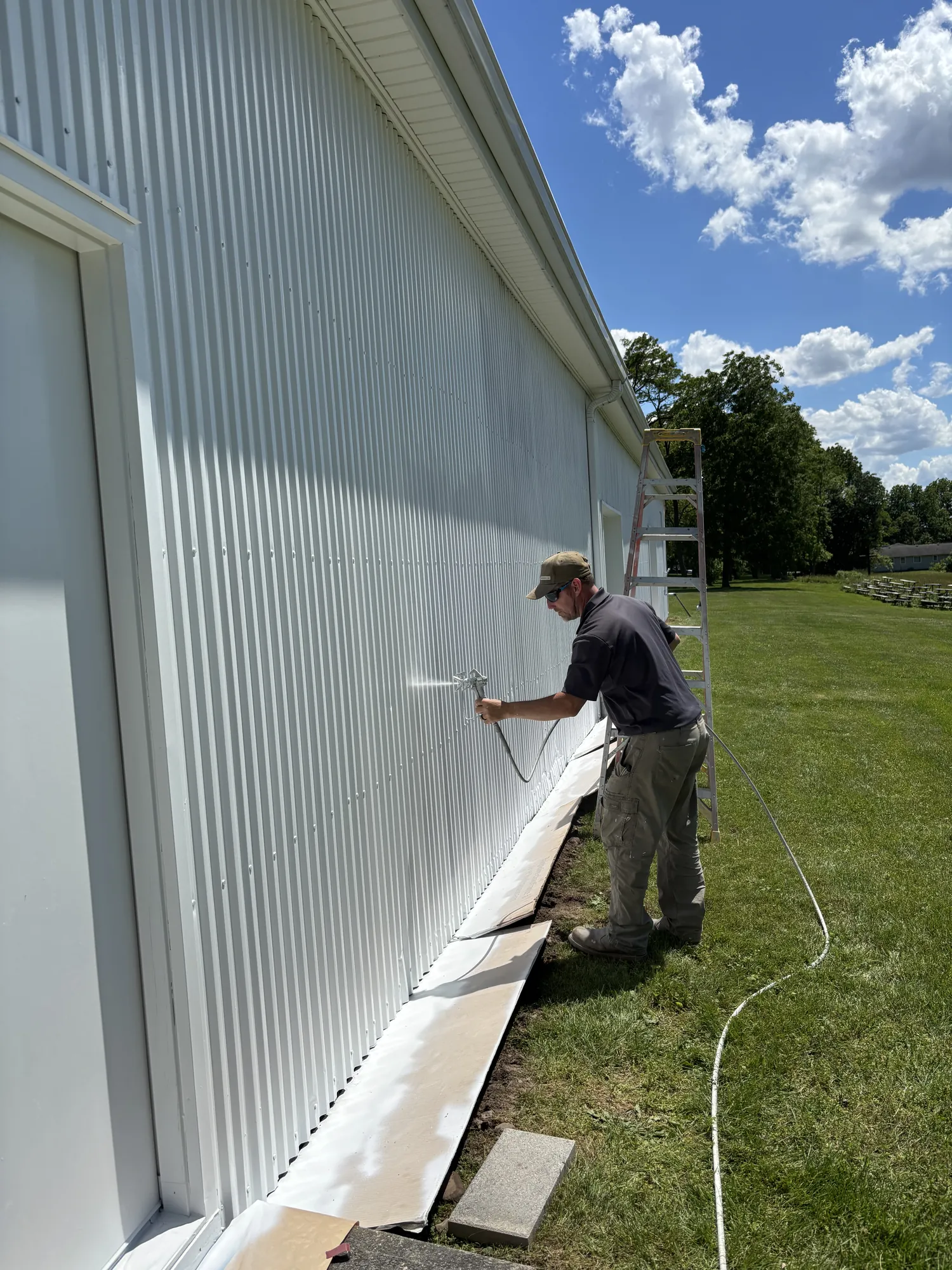 Painter sprays white coating on a corrugated metal wall; paper runners along the base protect the ground, ladder behind.