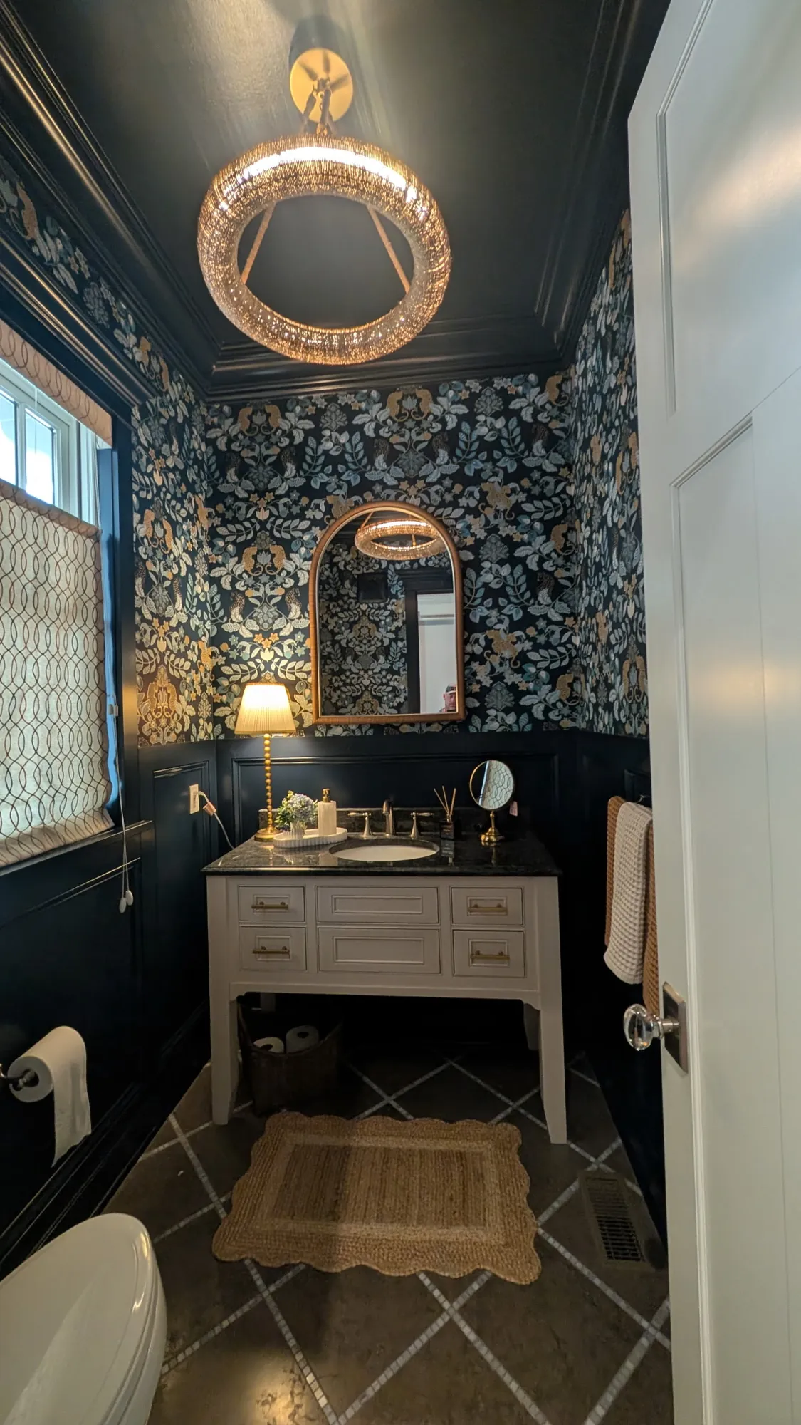 Powder room with dark navy wainscoting, crown and ceiling, leaf and leopard wallpaper, a ring chandelier and a white vanity