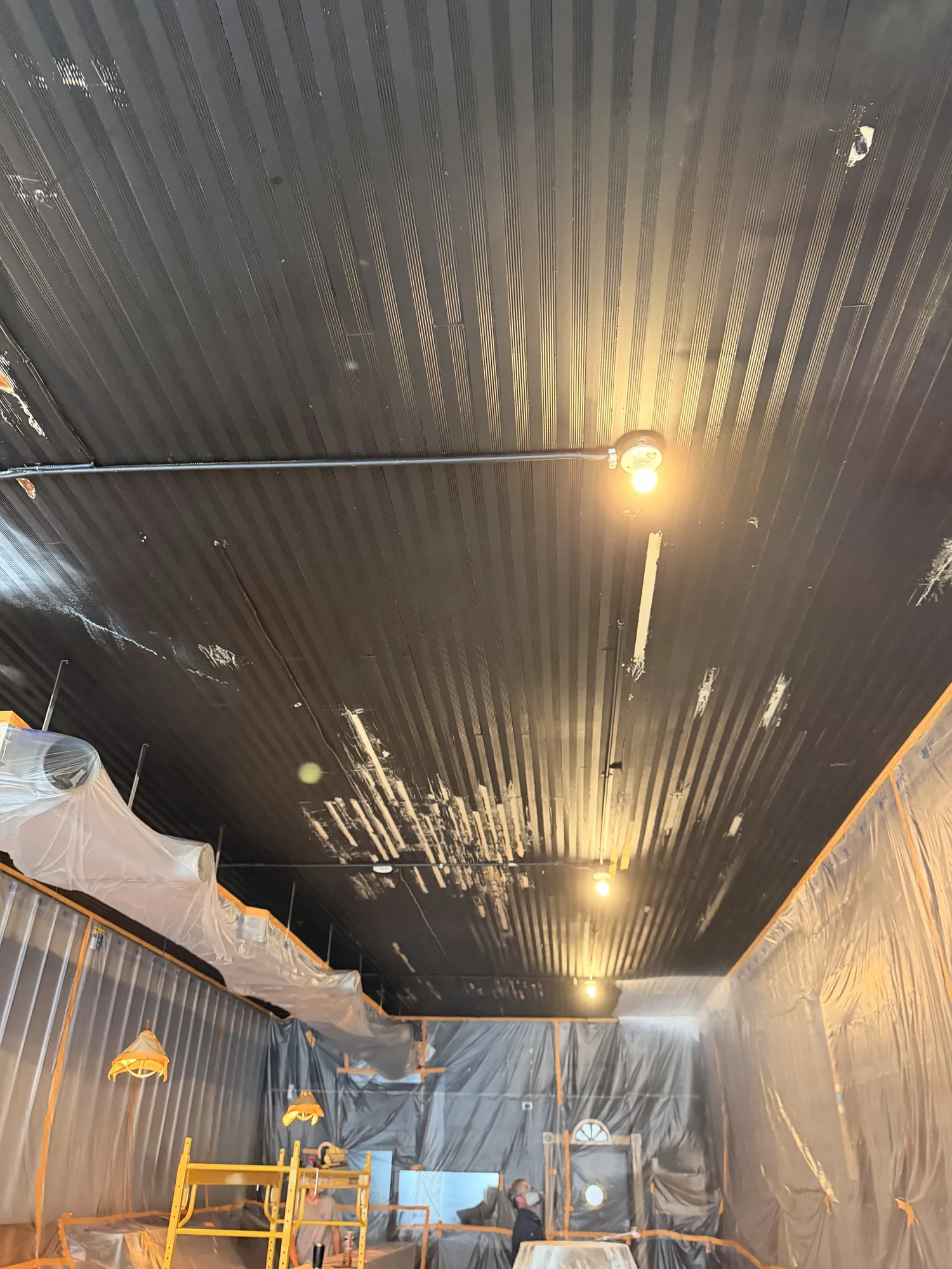 Looking up at a dark ribbed ceiling with chipped patches over walls, ductwork and light fixtures wrapped in plastic.