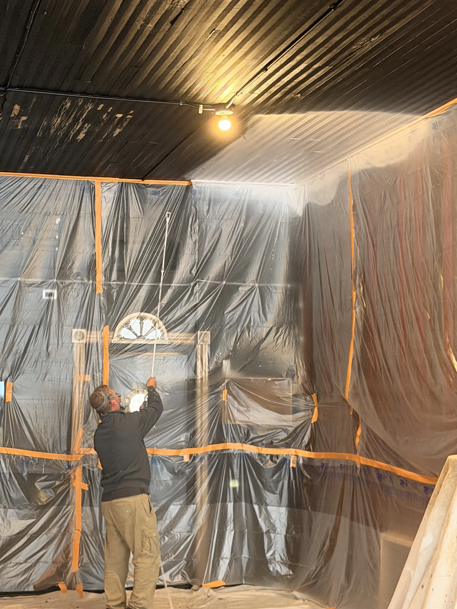 Painter in a respirator sprays a ceiling with an extension wand in a room sheeted in plastic and orange tape.